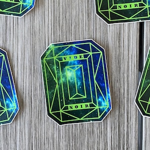 May include: Five green and blue stickers with a black outline and the words "VIDE NOIR" in white. The stickers are shaped like a gemstone and have a galaxy-like design.