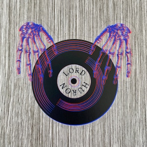May include: A black vinyl record with a red and blue 3D effect. The record has the words "LORD HURON" written on it. Two skeleton hands are holding the record.