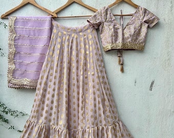 May include: A light purple and gold Indian bridal outfit with a skirt, a blouse, and a dupatta. The skirt has a ruffled hem and a gold print. The blouse has a V-neck and short sleeves. The dupatta is sheer and has a gold border.