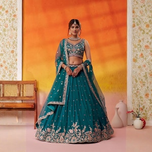 May include: A teal-colored, embroidered lehenga choli ensemble. The outfit features a heavily embellished bodice and a flowing skirt with floral embroidery. A matching dupatta and jewelry complete the look. The background is a gradient of orange and yellow.