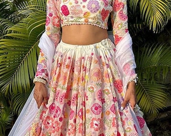 Può includere: Un lehenga bianco ricamato con un motivo floreale in tonalità di rosa, arancio, giallo e verde. Il lehenga è indossato da una persona con capelli lunghi e scuri e una fascia bianca. Il testo "Indian Wedding Outfit" è visibile nella parte inferiore dell'immagine.