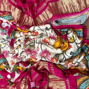 May include: A white and yellow floral patterned fabric with pink flowers and green leaves. The fabric is folded and draped over a pink and gold fabric with a fringe.