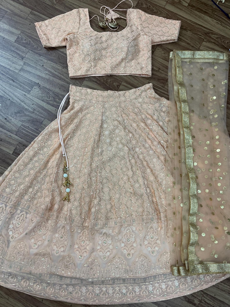 May include: A peach-colored Indian bridal outfit with a matching blouse, skirt, and sheer dupatta. The skirt and blouse are embroidered with silver sequins and beads, while the dupatta has gold sequins and a gold border.