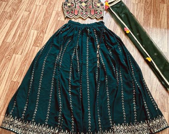 May include: A dark green skirt with gold embroidery and sequins. The skirt is paired with a matching crop top with intricate embroidery and a pink and red design. A sheer green dupatta with gold trim and tassels completes the outfit.
