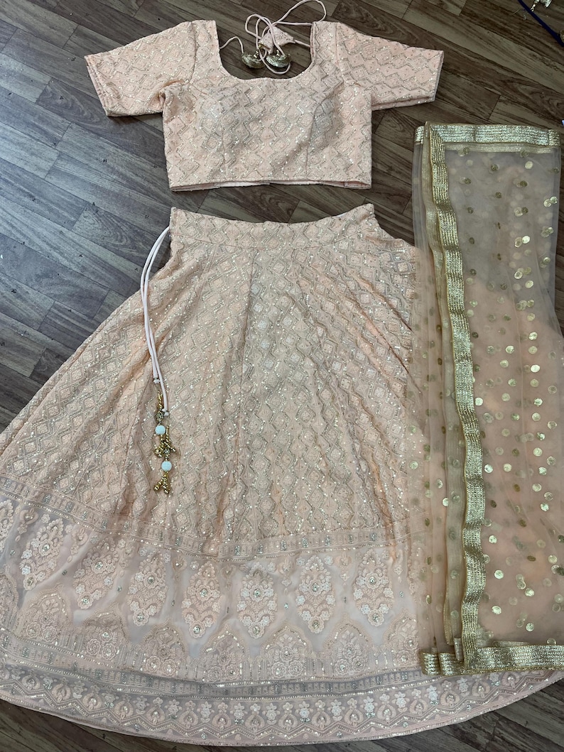 May include: A peach-colored Indian bridal outfit with a matching blouse, skirt, and sheer dupatta. The skirt and blouse are embroidered with gold sequins and the dupatta has gold sequins and a gold border.