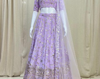 Può includere: Un completo formale in due pezzi color lavanda con intricati abbellimenti dorati. Il set include un top corto, una gonna lunga e una sciarpa trasparente e fluida. L'outfit è esposto su un manichino.