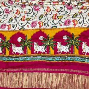 May include: A white fabric with a floral pattern of pink and orange lotus flowers and green leaves. The bottom border features a repeating pattern of white cows walking under palm trees with yellow, orange, pink, and blue accents.