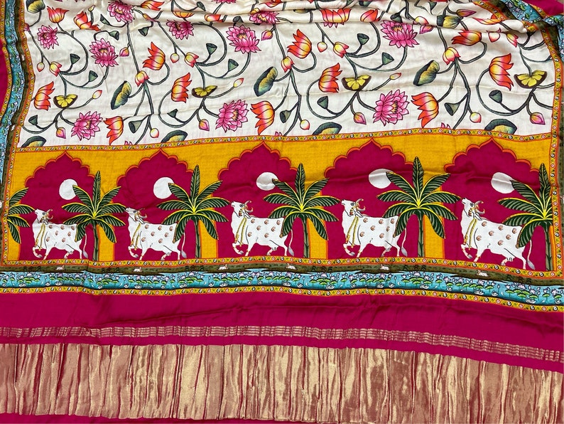 May include: A white fabric with a floral pattern of pink and orange lotus flowers and green leaves. The bottom border features a repeating pattern of white cows walking under palm trees with yellow, orange, pink, and blue accents.