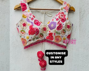 以下が含まれることがあります： ピンクとマルチカラーの花柄が刺繍された白いクロップトップ。トップはVネックで、ピンクのビーズで縁取られています。画像には「CUSTOMISE IN ANY STYLES」というテキストが表示されています。