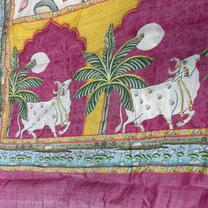May include: A pink and yellow fabric with a white background. The fabric features a repeating pattern of cows, palm trees, and lotus flowers. The fabric is a traditional Indian design.
