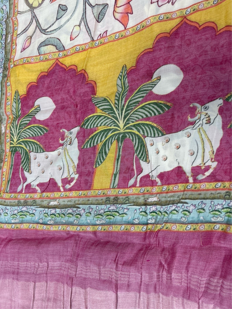 May include: A pink and yellow fabric with a white background. The fabric features a repeating pattern of cows, palm trees, and lotus flowers. The fabric is a traditional Indian design.