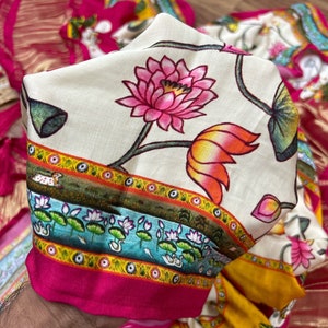 May include: A white fabric with a pink and orange floral pattern. The fabric has a border with a blue, yellow, and green design. The fabric is folded in half and held by a hand.