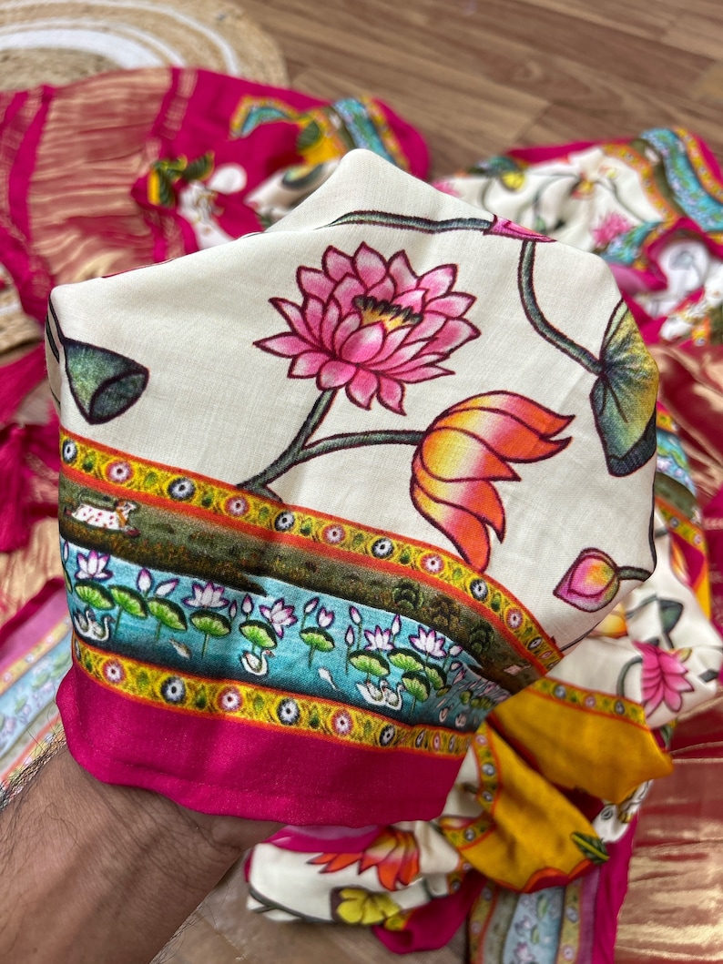 May include: A white fabric with a pink and orange floral pattern. The fabric has a border with a blue, yellow, and green design. The fabric is folded in half and held by a hand.