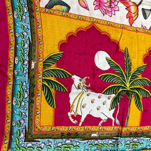 May include: A pink fabric with a colorful design featuring a white cow, palm trees, and a moon. The design is framed by a yellow, orange, and blue border with floral motifs.