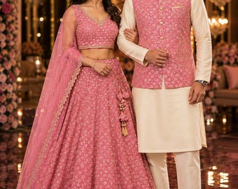May include: A pink lehenga choli with intricate gold embroidery and a matching sheer dupatta, paired with a cream-colored kurta and a pink embroidered Nehru jacket featuring gold buttons and a mandarin collar, creating a coordinated ensemble.