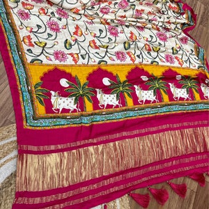 May include: A white silk saree with a floral print and a pink and gold border. The saree has a traditional Indian design with cows and palm trees.