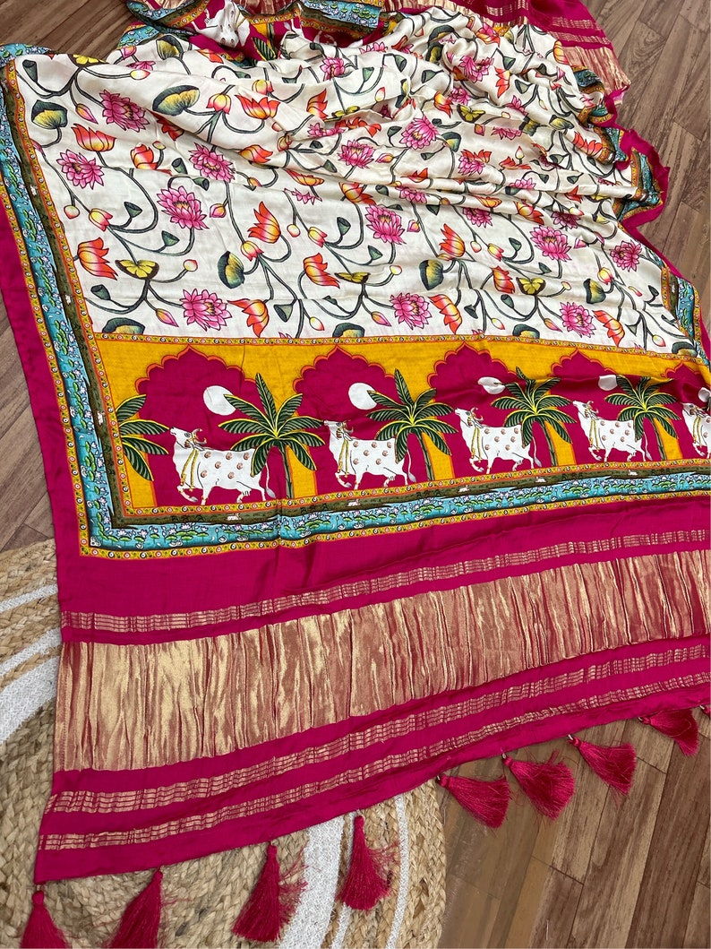 May include: A white silk saree with a floral print and a pink and gold border. The saree has a traditional Indian design with cows and palm trees.
