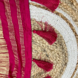 May include: A close-up of a pink fabric with gold stripes and a fringe of red tassels. The fabric is lying on a woven rug.