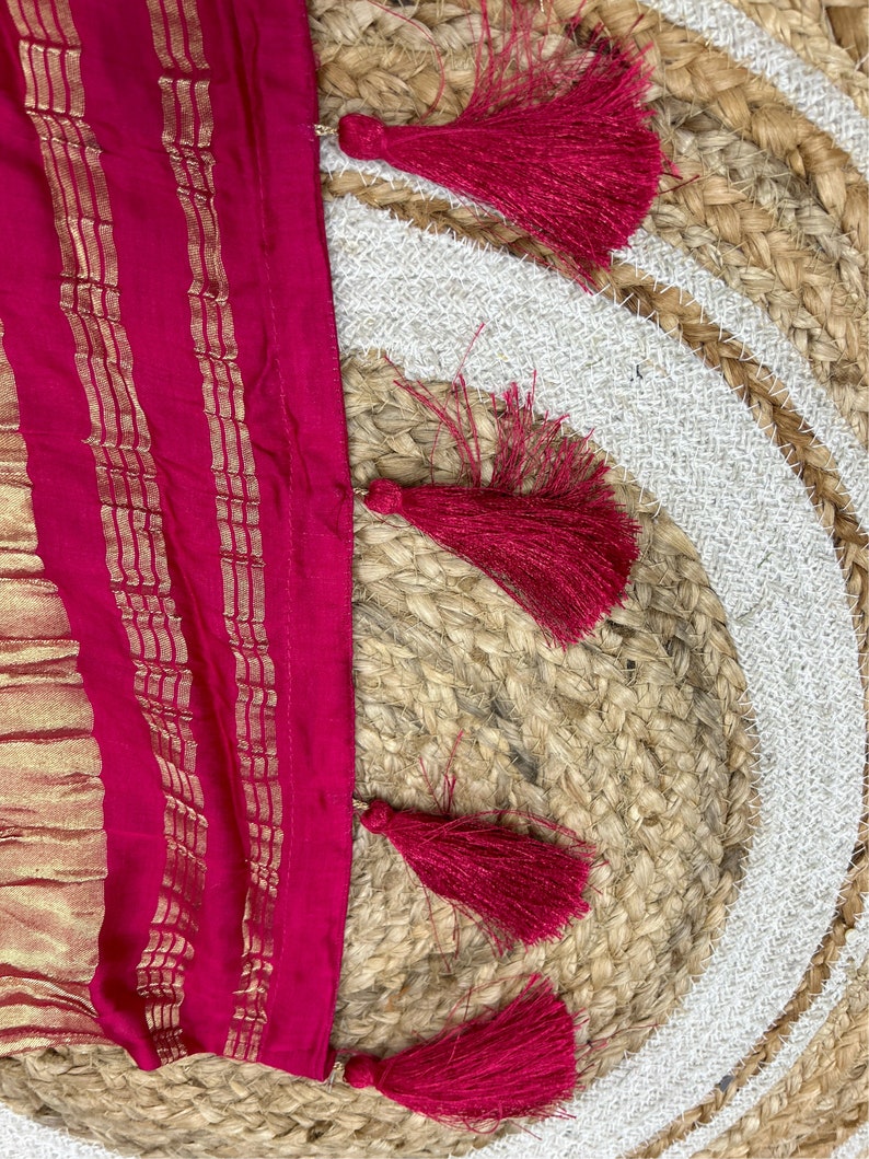 May include: A close-up of a pink fabric with gold stripes and a fringe of red tassels. The fabric is lying on a woven rug.