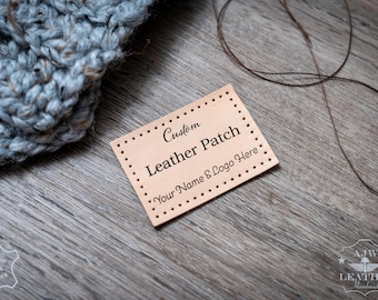 Può includere: Una toppa rettangolare in pelle color cuoio con fori pre-perforati lungo i bordi. Il testo stampato sulla toppa recita Custom Leather Patch Your Name & Logo Here. La toppa è posizionata vicino a un tessuto lavorato a maglia grigio e un filo marrone.