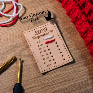 Op de afbeelding: Een rechthoekig lichtbruin leren label met de gegraveerde tekst 2023 Temperature °C en een lijst met temperatuurbereiken van <6 tot 25+. Het label heeft kleine gaatjes voor garenmonsters, met wit en rood garen door de bovenste rij geregen.
