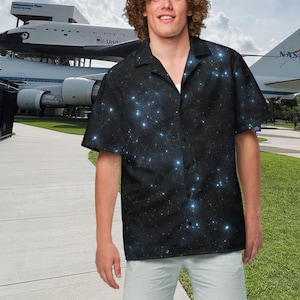 May include: Black button-down shirt with a celestial galaxy print featuring blue and white stars. The short-sleeved shirt has a classic collar and is worn with light-colored shorts. The shirt is made of a lightweight fabric.