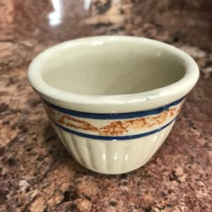 May include: A small, white ceramic cup with a blue and brown rim. The cup has a fluted design and is perfect for holding small items.