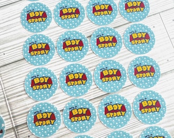 May include: A collection of round stickers with the text "It's a Boy Story" in red and yellow on a light blue background with white triangle patterns. The stickers are arranged on a wooden surface.