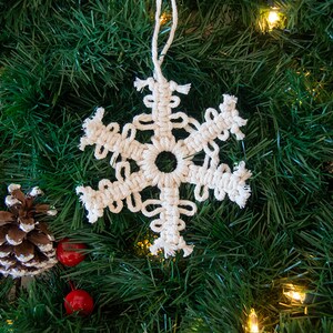 May include: A white macrame snowflake ornament hanging on a green artificial Christmas tree with brown pine cones and red berries.