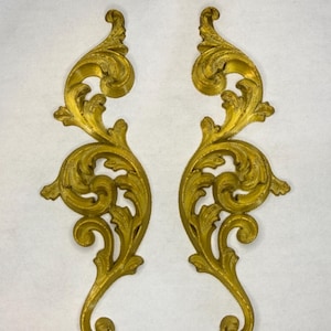 May include: Two ornate, gold-colored decorative scrollwork pieces. Each piece features a symmetrical design with swirling leaves and a curved, elongated bottom. The pieces are likely made of metal and are suitable for home decor.