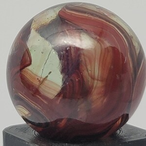 May include: A red, white, and brown swirl glass marble. The marble has a glossy finish and is sitting on a black stand.