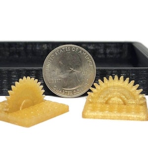 May include: Two small, light-yellow, 3D-printed objects resembling a saw and a sun, each on a square base. A U.S. quarter coin is next to the objects for size comparison. A black, rectangular tray is in the background.