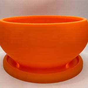 May include: Bright orange, rounded planter pot with a matching saucer base. The pot has a smooth, matte finish and a wide opening. A modern, decorative item for indoor or outdoor use.