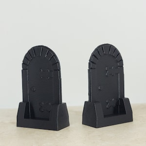 May include: Two black miniature tombstone decorations. Each tombstone has an arched top with vertical lines, a rectangular body, and a base. The tombstones have a cross and bone design. Perfect for Halloween or spooky decor.