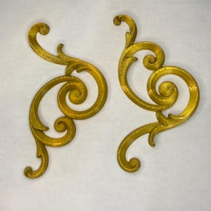 May include: Two ornate, gold-colored decorative scrollwork pieces. Each piece features a swirling, symmetrical design with rounded edges and a textured surface. The pieces are likely for furniture or architectural embellishment.