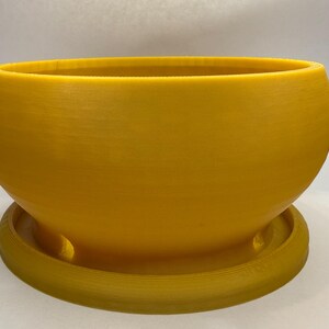 May include: A yellow, rounded planter with a matching saucer. The planter has a smooth, matte finish and a wide opening. The saucer has a raised edge and three drainage holes. This decorative planter is suitable for indoor or outdoor use.