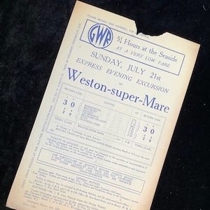 Puede incluir: Un cartel vintage de Great Western Railway (GWR) que anuncia una excursión de un día a Weston-super-Mare el domingo 21 de julio. El cartel presenta un logotipo azul y blanco con las iniciales GWR y un horario de trenes con las horas de salida y las tarifas para varias estaciones.