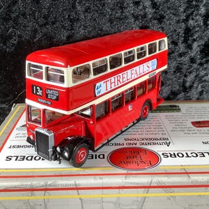 May include: A red and white double-decker bus model with the text "THRELFALL'S" on the side. The model bus has black tires and a silver front grill. The bus is a collectible item.