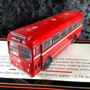 May include: A red model bus with white detailing, marked "EASTERN COUNTIES THORPE STN 90". The miniature bus is a 1:76 scale die-cast model, with black wheels. It is a replica of a classic bus.