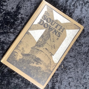 Op de afbeelding: Een vintage boek getiteld "The South Downs" met een illustratie van een windmolen op de omslag. De omslag is lichtbruin met zwart-witte afbeeldingen. Het boek staat schuin.