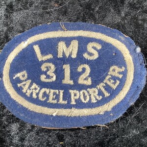 Peut inclure: Un écusson ovale en tissu bleu avec des lettres brodées en beige indiquant LMS 312 PARCEL PORTER. Le texte est disposé avec LMS en haut, 312 au centre et PARCEL PORTER incurvé le long du bord inférieur de l'écusson en tissu.
