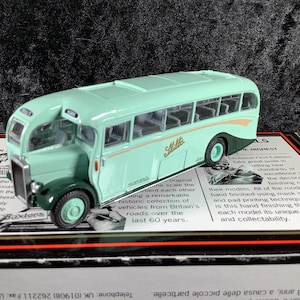 Könnte beinhalten: Ein mintgrüner und dunkelgrüner Modellbus mit dem Namen "Mills" an der Seite. Der Bus im Vintage-Stil hat detaillierte Fenster und Räder. Er wird auf einer Box mit Text präsentiert.
