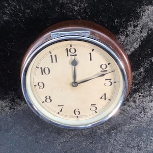 May include: A circular clock with a cream face, black numerals, and black hands. It is set within a silver metal bezel and a dark brown wooden frame. The clock displays the time as approximately 2:10.