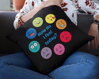 May include: A black throw pillow with a variety of colorful emojis and the text "How do I feel today?" printed on it. The pillow is being held by a person sitting on a gray couch.