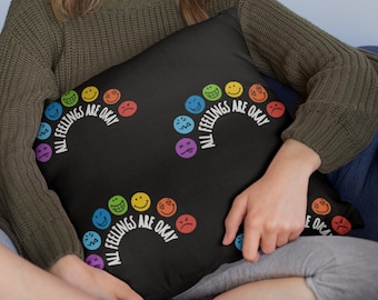 May include: A black throw pillow with a rainbow of colorful smiley faces and the text "All Feelings Are Okay".