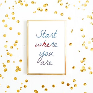 Peut inclure: Une impression encadr&eacute;e avec les mots "Start where you are" en lettrage aquarelle bleu et rouge. Le cadre dor&eacute; repose sur une surface blanche parsem&eacute;e de paillettes dor&eacute;es, cr&eacute;ant une pi&egrave;ce d&eacute;corative et inspirante.