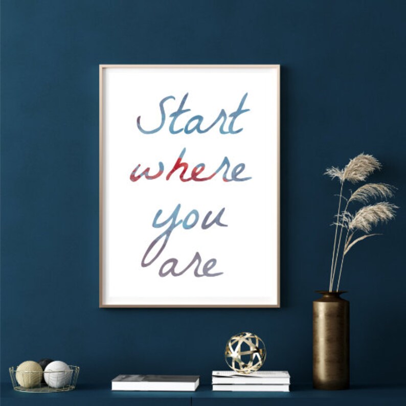 Peut inclure: Une affiche encadr&eacute;e avec l'inscription "Start where you are" en lettres aquarelles bleues et rouges. L'affiche est accroch&eacute;e &agrave; un mur bleu fonc&eacute;, avec des objets d&eacute;coratifs sur une &eacute;tag&egrave;re en dessous.
