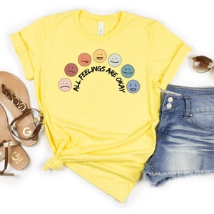 Op de afbeelding: Geel T-shirt met een regenboog van verschillende gekleurde gezichten en de tekst "Alle gevoelens zijn ok&eacute;".