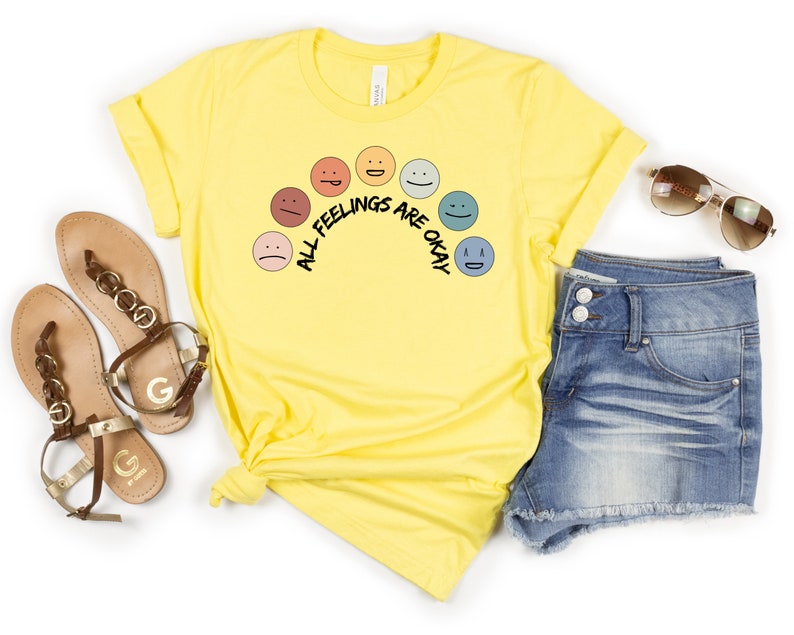 Op de afbeelding: Geel T-shirt met een regenboog van verschillende gekleurde gezichten en de tekst "Alle gevoelens zijn ok&eacute;".