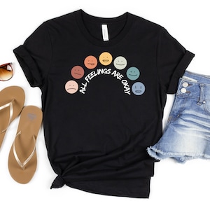 Op de afbeelding: Zwart T-shirt met een regenboog van kleurrijke gezichten en de tekst "Alle gevoelens zijn ok&eacute;".